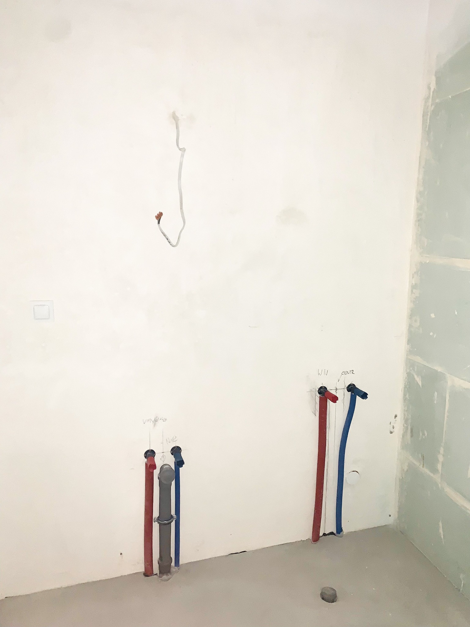 Surowa ściana w trakcie prac przygotowawczych w łazience: widoczne wystające rury wodne (czerwone i niebieskie) z opisami, rura kanalizacyjna oraz fragment ściany pokryty płytami kartonowo-gipsowymi.