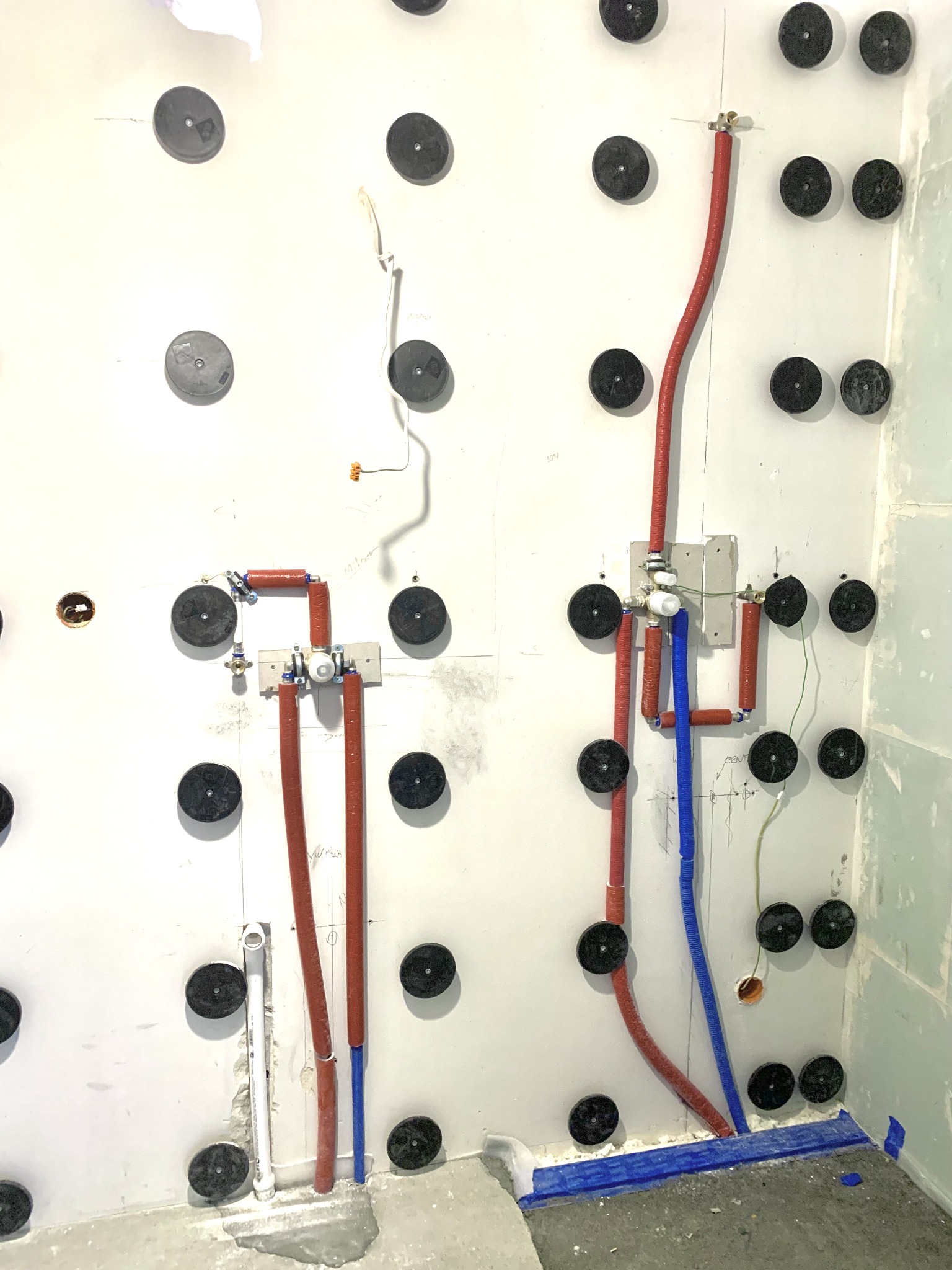 Ściana w trakcie przygotowania do wykończenia łazienki: widoczne rury, przewody, zamocowania, zabezpieczenia i oznaczenia na płycie kartonowo-gipsowej.