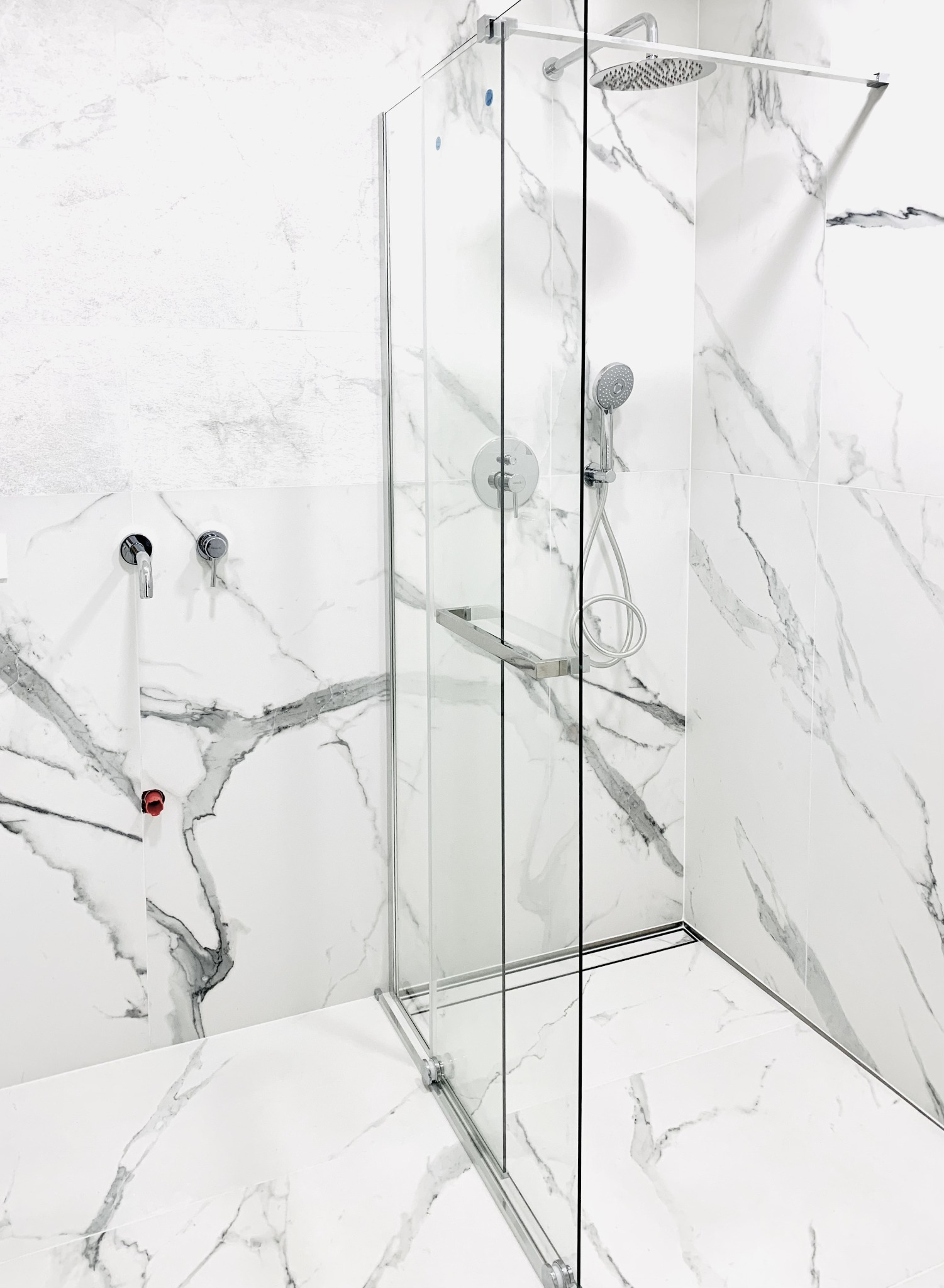 Nowoczesna kabina prysznicowa z chromowanymi elementami, osadzona na białych płytkach imitujących marmur, z widocznymi bateriami i słuchawką prysznicową.