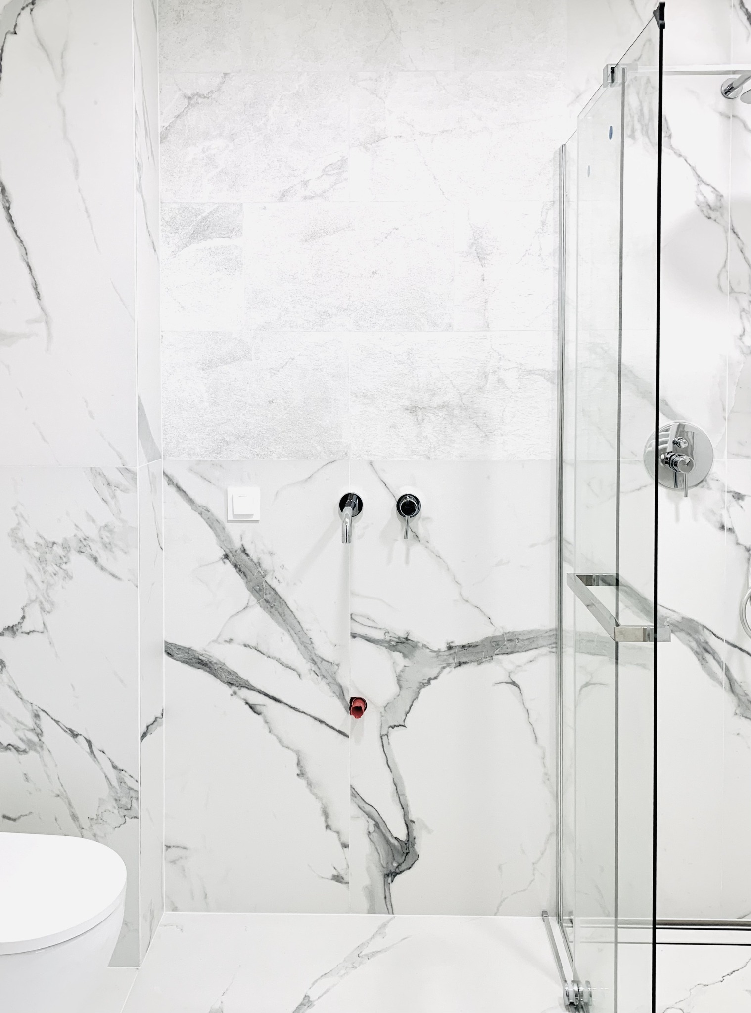 Nowoczesna łazienka z białymi płytkami imitującymi marmur, szklana kabina prysznicowa z chromowanym uchwytem, minimalistyczna armatura i częściowo widoczna biała toaleta.