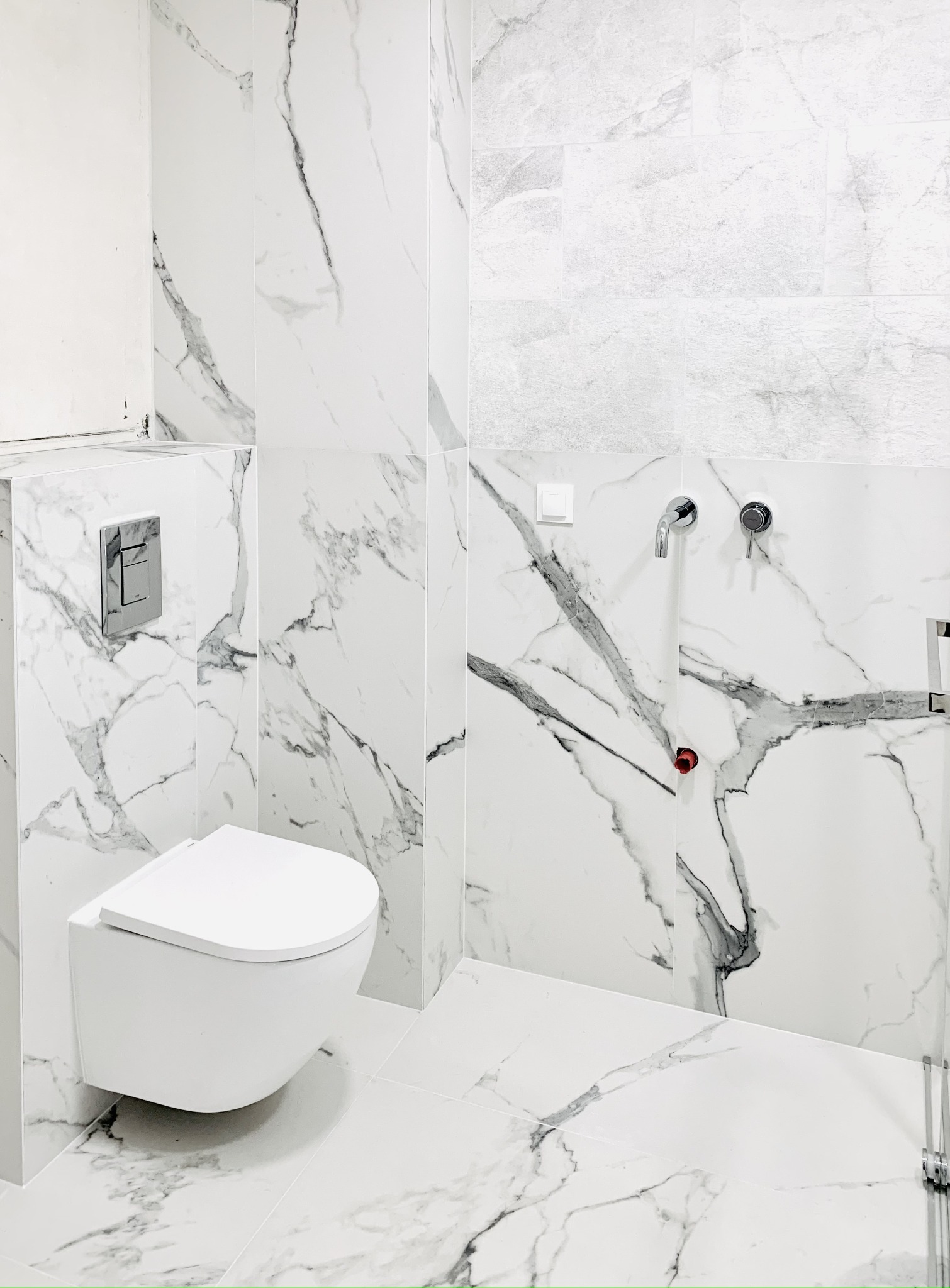 Nowoczesna łazienka wykończona białymi płytkami imitującymi marmur, z podwieszaną toaletą i chromowaną armaturą. Fragment ściany z szarą, strukturalną płytką. Widoczne niedokończone prace przy...