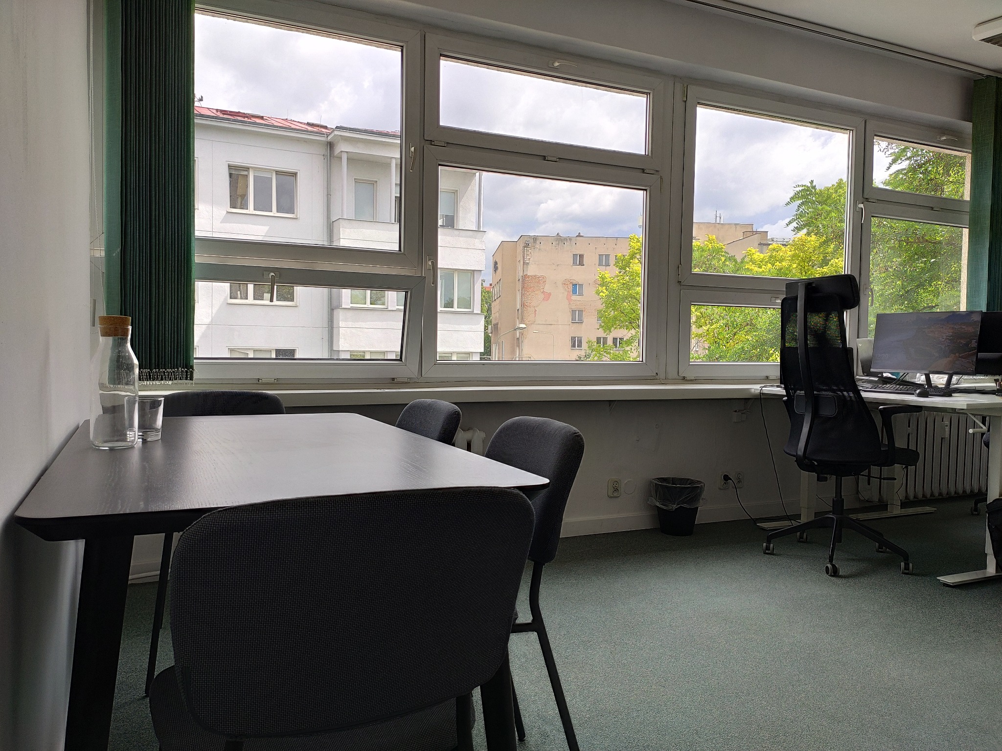 Jasne biuro w Poznaniu z dużym oknem wychodzącym na budynki i drzewa. Widać stół konferencyjny z krzesłami, biurko z komputerem i krzesłem obrotowym. Na stole szklana butelka z wodą i szklanki.