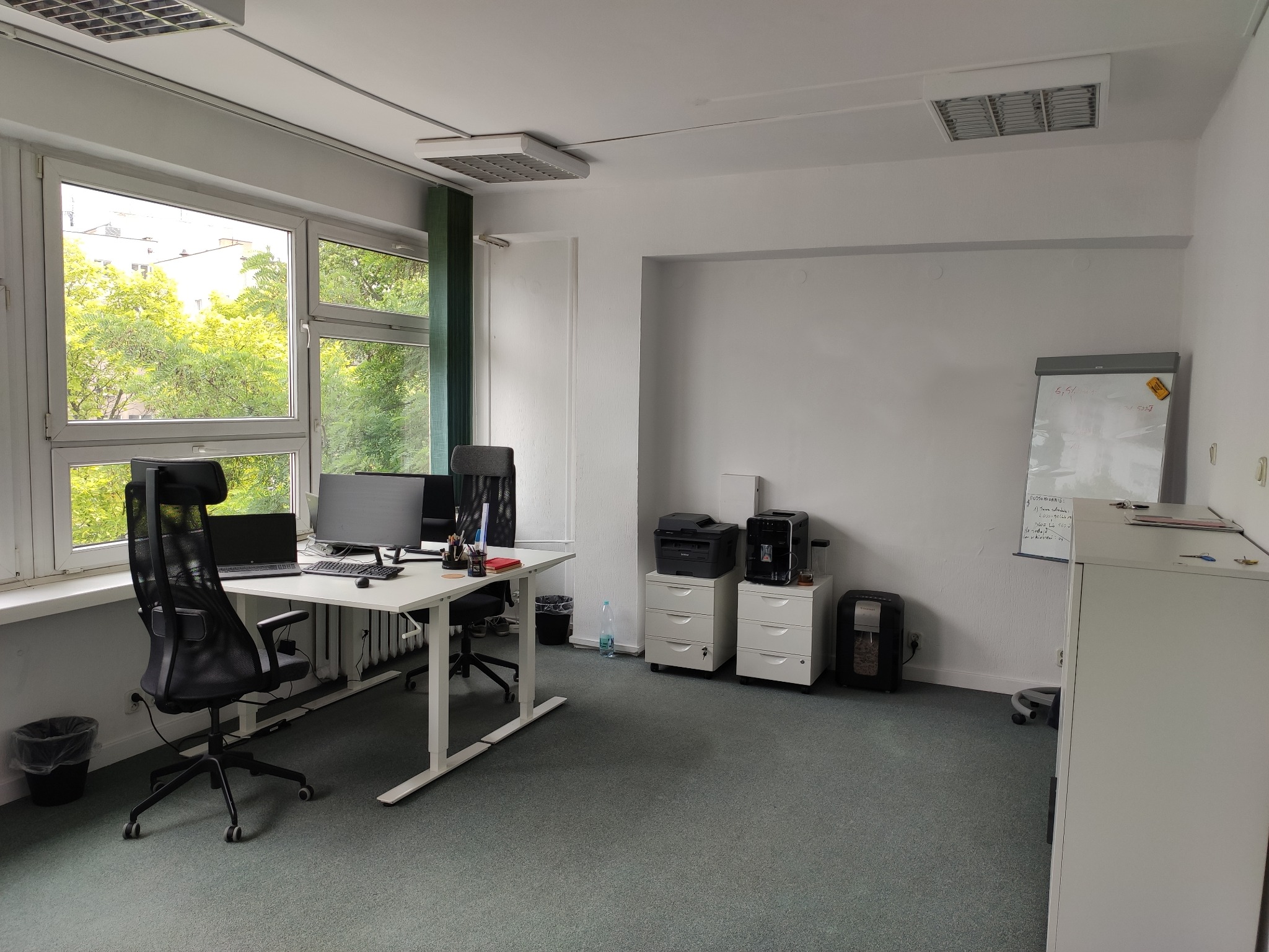Jasne biuro z dwoma biurkami, krzesłami ergonomicznymi, komputerami, szafkami, drukarką i flipchartem, widok z okna na zieleń drzew.