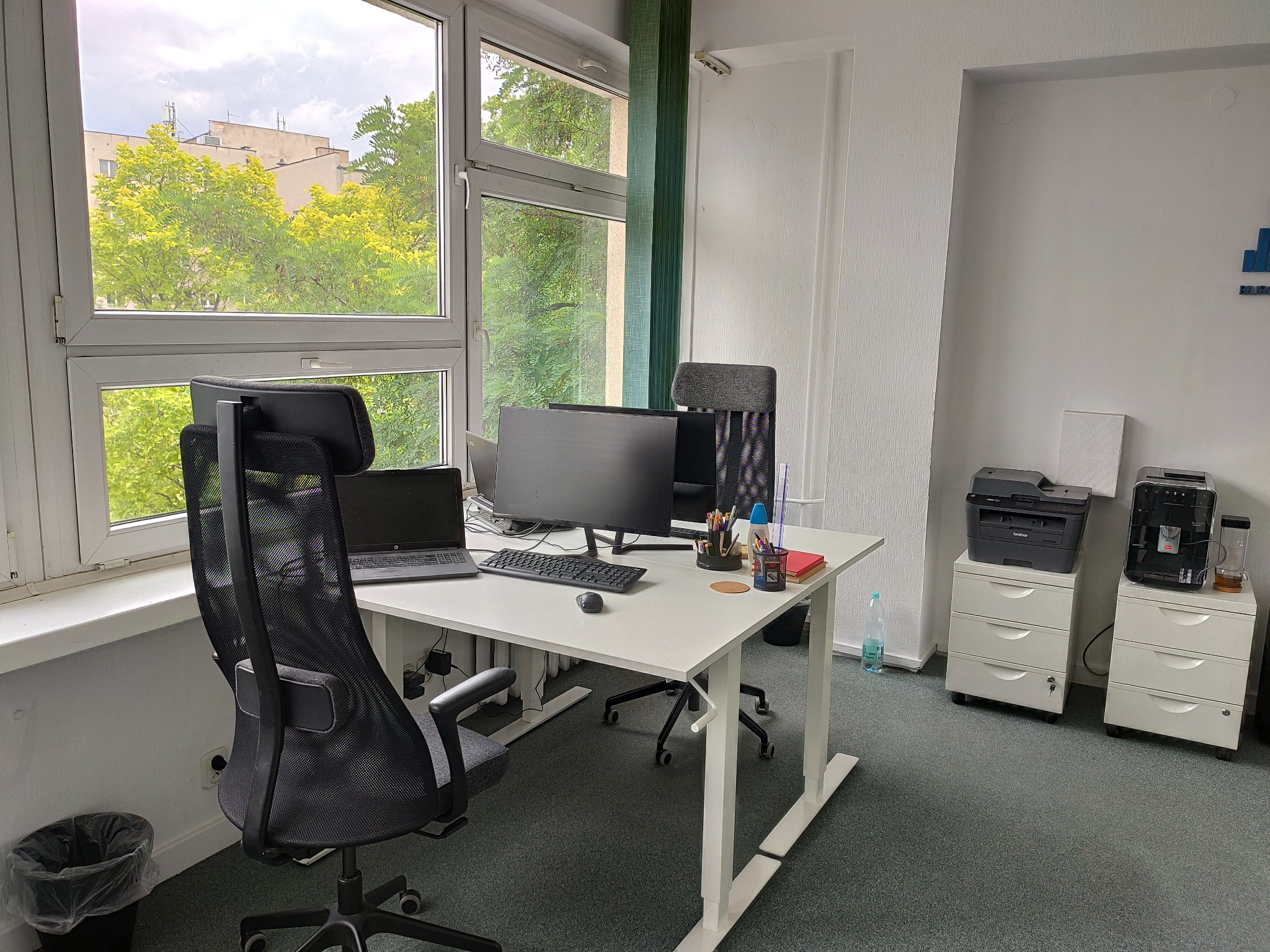 Jasne biuro z regulowanym biurkiem, krzesłem biurowym z siatki, laptopem i monitorem na biurku, drukarką i ekspresem do kawy na szafkach z szufladami. Widok z okna na zieleń.
