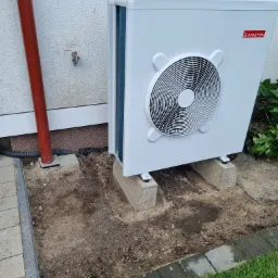 Biała pompa ciepła marki Ariston, zamontowana na betonowych bloczkach przy ścianie budynku z widoczną rurą spustową i zaworem.