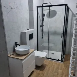 Nowoczesna łazienka z kabiną prysznicową typu walk-in z czarnymi profilami, białą umywalką nablatową na szafce z drewnianym blatem, wiszącą toaletą i grzejnikiem drabinkowym. Na ścianie widoczne...