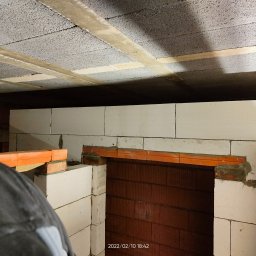 FRAAN-DOM Dominik Dryka - Wnętrze w trakcie budowy: sufit z betonowych bloków, ściany z białych bloczków, widoczny otwór z ceglaną obudową, fragment osoby w lewym dolnym rogu kadru.