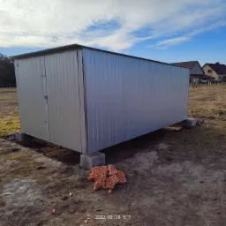 Metalowy garaż blaszany na betonowych bloczkach, świeżo postawiony na działce, widoczne porozrzucane cegły, w tle domy i drzewa.