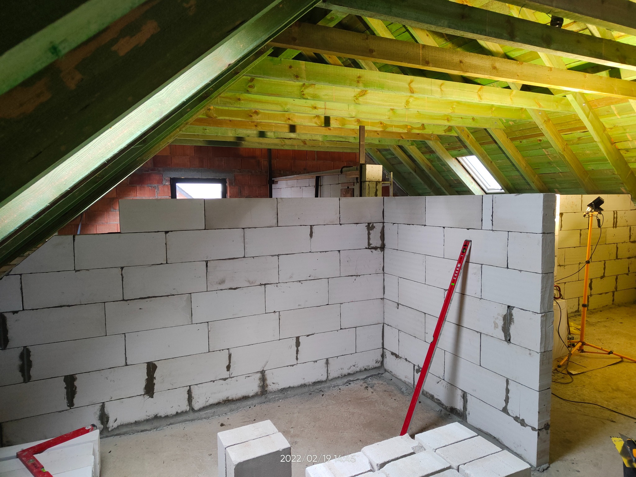 Wnętrze poddasza w trakcie budowy ścian z białych bloczków, widoczna drewniana konstrukcja dachu, czerwona poziomica oparta o ścianę, lampa budowlana oświetlająca wnętrze.