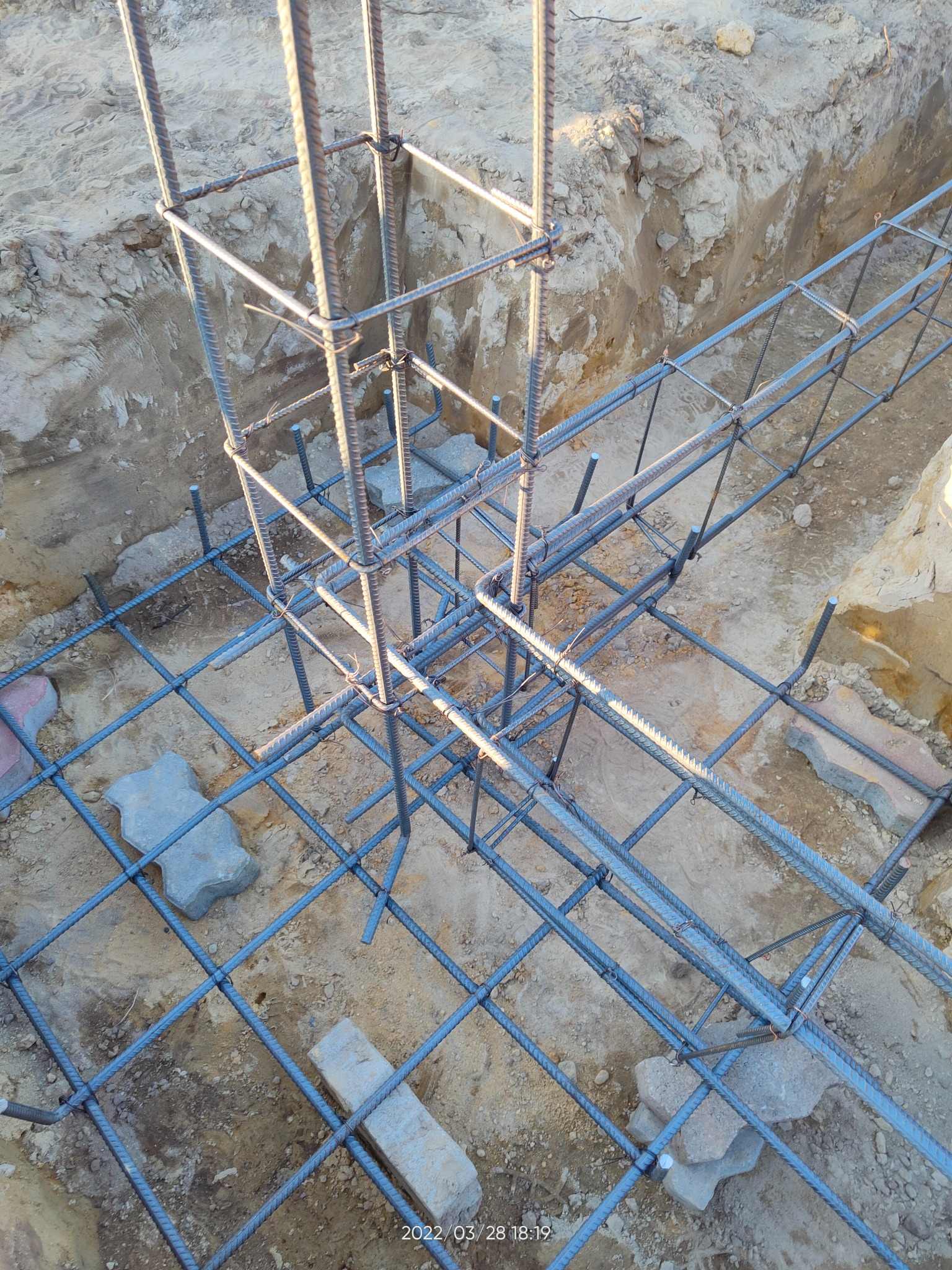 Zbrojenie fundamentów z prętów stalowych, widoczne w wykopie, z elementami betonowej kostki brukowej na dnie.