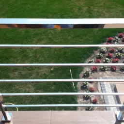 Błyszcząca, nierdzewna balustrada balkonowa z poziomymi prętami, widok z góry na zadbany trawnik i rabatę kwiatową z nawodnieniem kropelkowym.