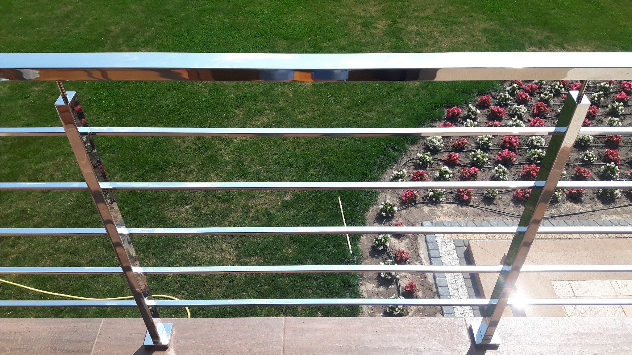 Błyszcząca, nierdzewna balustrada balkonowa z poziomymi prętami, widok z góry na zadbany trawnik i rabatę kwiatową z nawodnieniem kropelkowym.