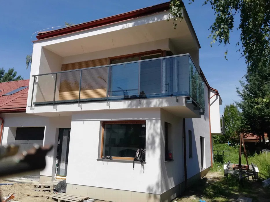 Nowoczesny dom z balkonem zabezpieczonym szklaną balustradą z metalowymi elementami, widok z zewnątrz, w tle drzewa i niebo.