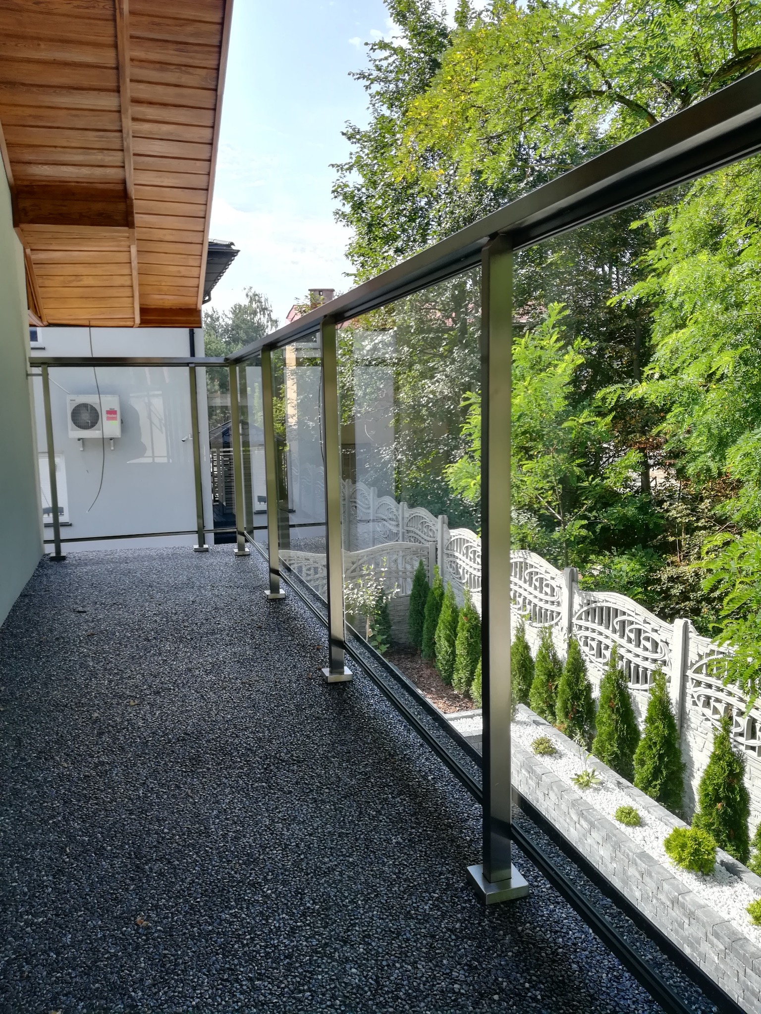 Nowoczesny balkon z szarą, kamienną posadzką i szklaną balustradą z ciemnymi, metalowymi ramami, widok na ogród z tujami i dekoracyjnym, białym ogrodzeniem