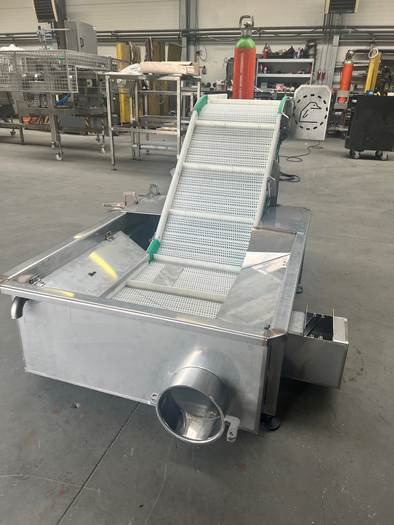 Metalowa konstrukcja z taśmą transportującą, prawdopodobnie element linii produkcyjnej, widoczne spawane łączenia, obudowa ze stali nierdzewnej, w tle butle z gazem.