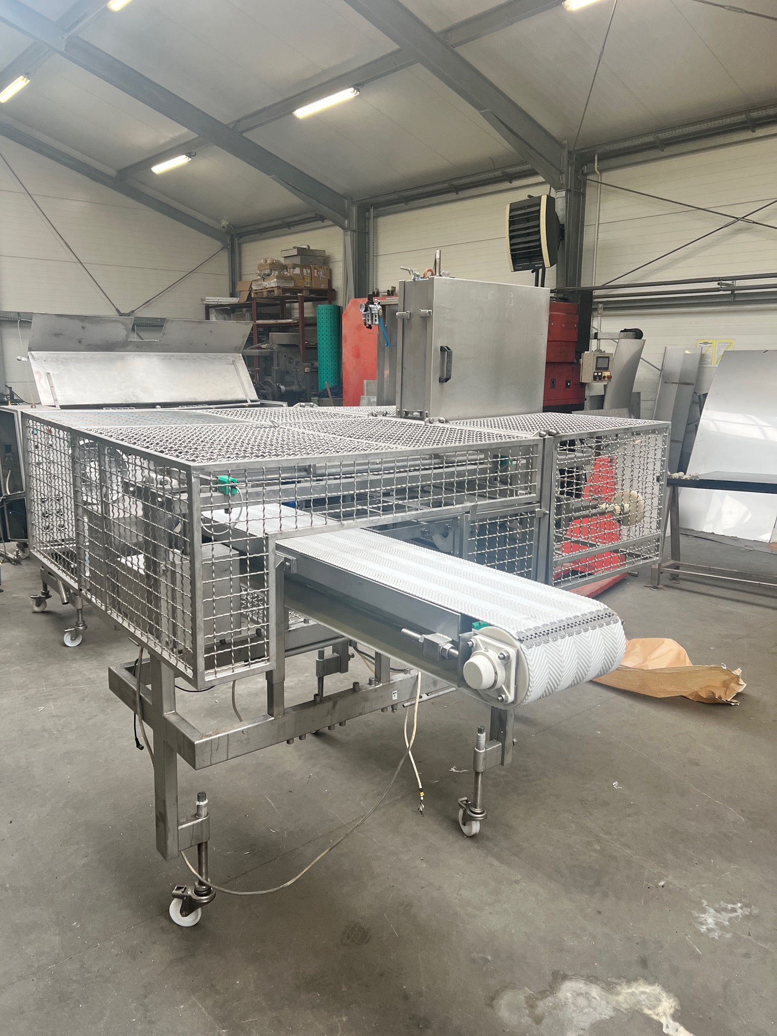 Konstrukcja ze stali nierdzewnej z siatką i taśmociągiem w hali produkcyjnej, widoczne elementy sterujące i inne maszyny w tle.
