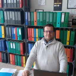 Mężczyzna w okularach siedzi za biurkiem z laptopem, otoczony regałami pełnymi segregatorów i ramkami z certyfikatami na ścianie.