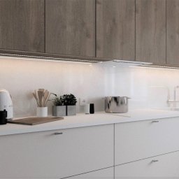 Home by M Martyna Matuszak - Nowoczesna kuchnia z białymi szafkami dolnymi i drewnopodobnymi górnymi, podświetlona listwą LED, z ekspresem do kawy, czajnikiem SMEG i garnkiem na blacie