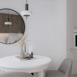 Home by M Martyna Matuszak - Jasne wnętrze kuchni z okrągłym lustrem, białym stołem i szarymi krzesłami, widok na zabudowę kuchenną z piekarnikiem i lodówką.