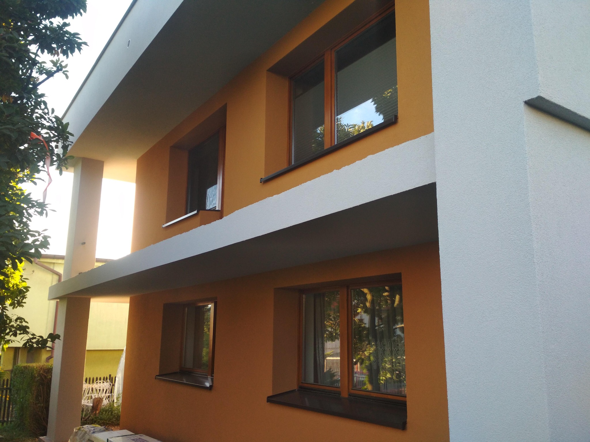 Dwukondygnacyjny dom jednorodzinny po częściowym nałożeniu elewacji w kolorze pomarańczowym i białym, z widocznymi oknami z brązowymi ramami i szarymi elementami architektonicznymi.