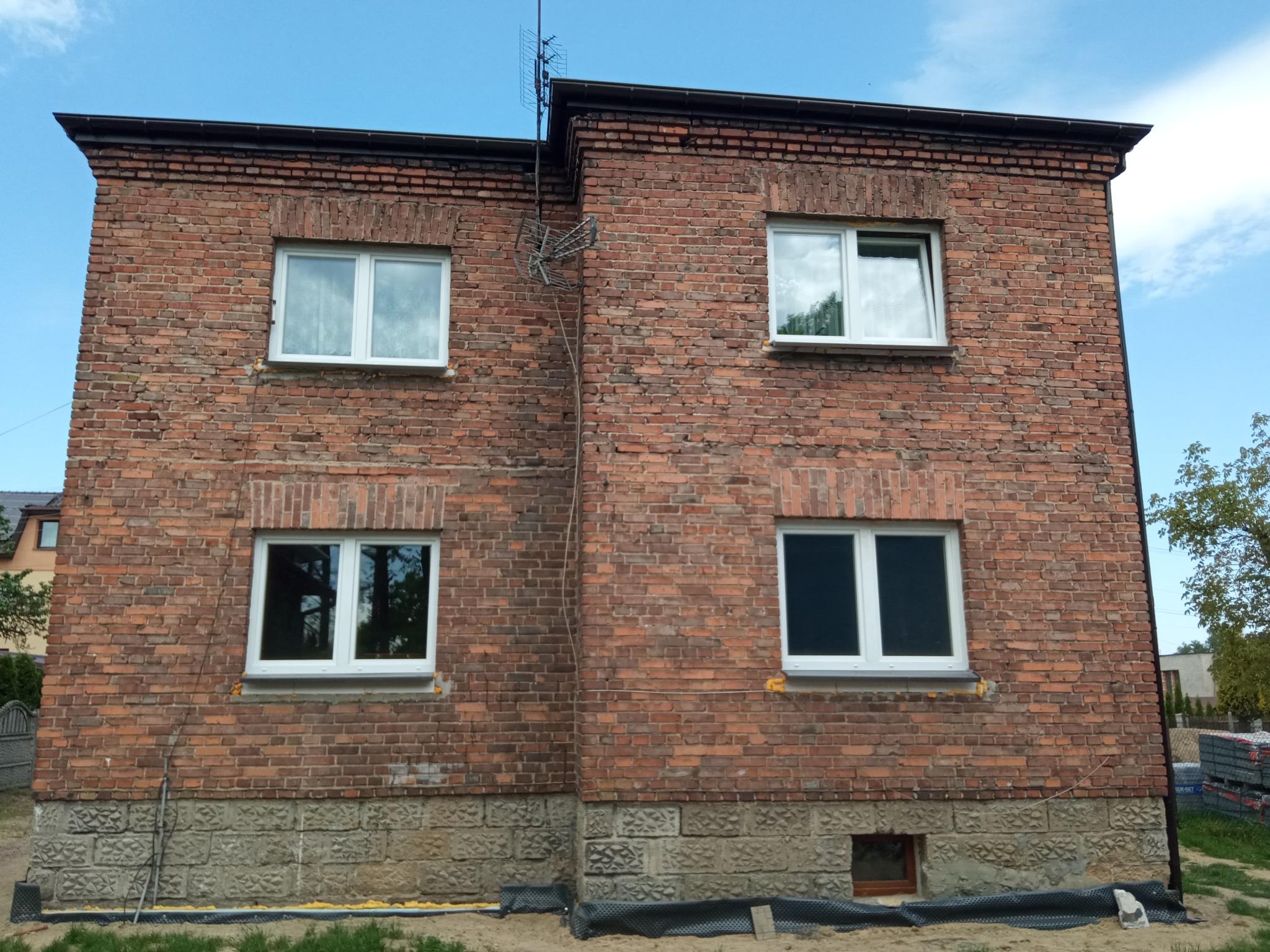 Dwukondygnacyjny budynek z czerwonej cegły z nowymi białymi oknami, widoczna antena na dachu i przygotowany teren wokół fundamentów z czarną folią.