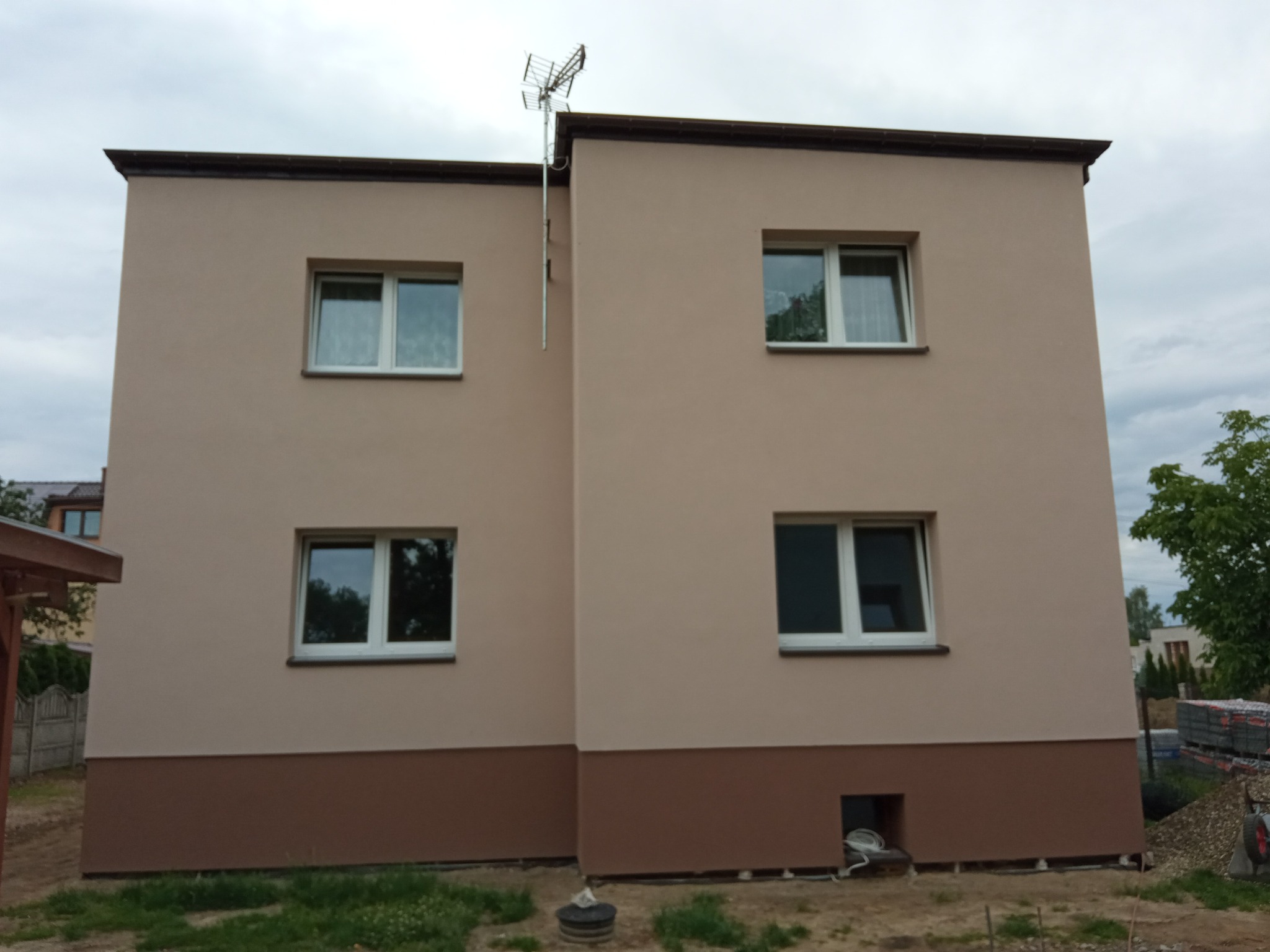 Dwukondygnacyjny dom po ociepleniu elewacji w odcieniach beżu i brązu, z widoczną anteną telewizyjną na dachu i stosem materiałów budowlanych obok.