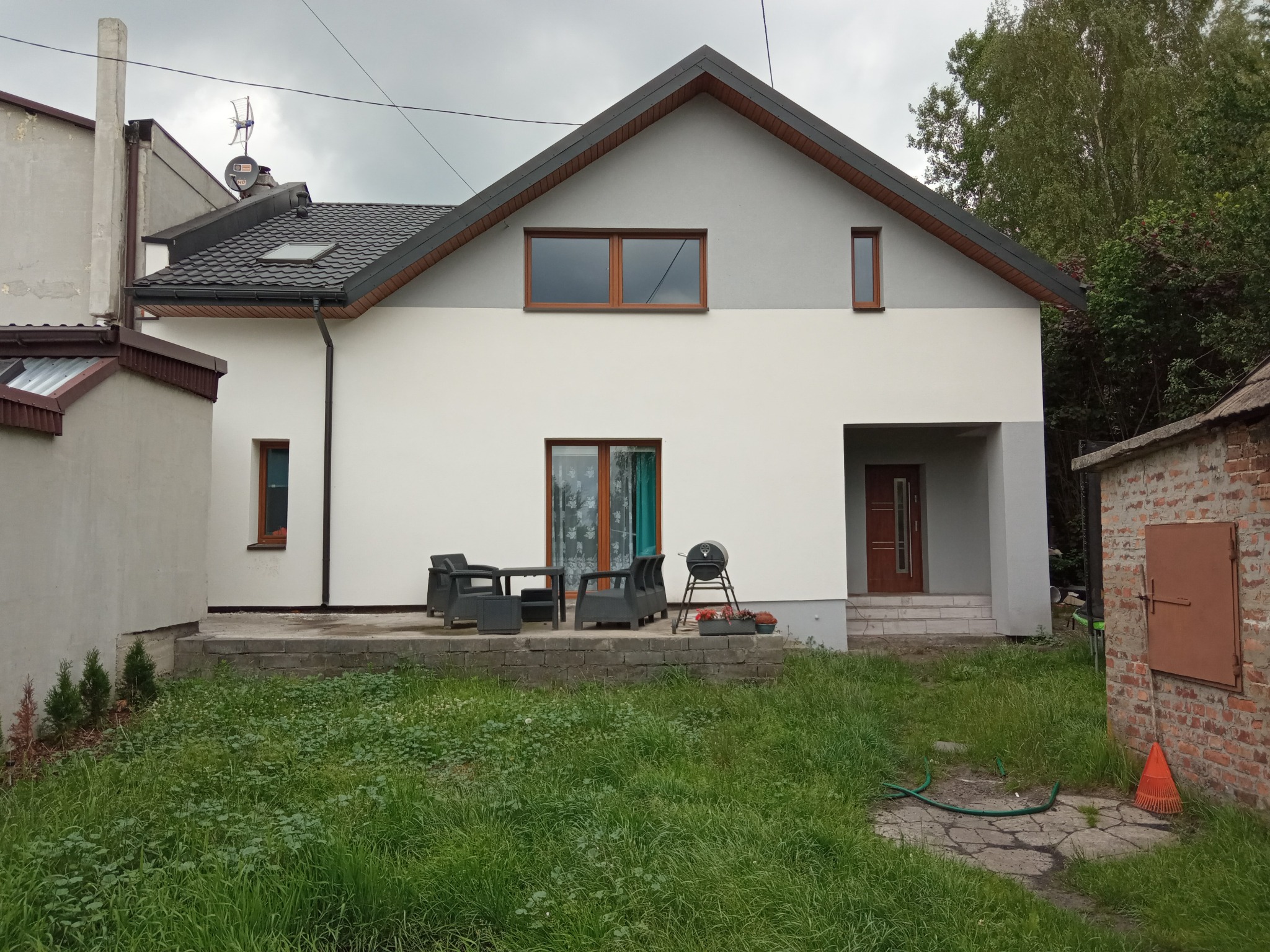 Ocieplony dom jednorodzinny w Sosnowcu z wykończoną elewacją w kolorze białym i szarym, widok na taras z meblami ogrodowymi i grillem, trawnik z wężem ogrodowym.