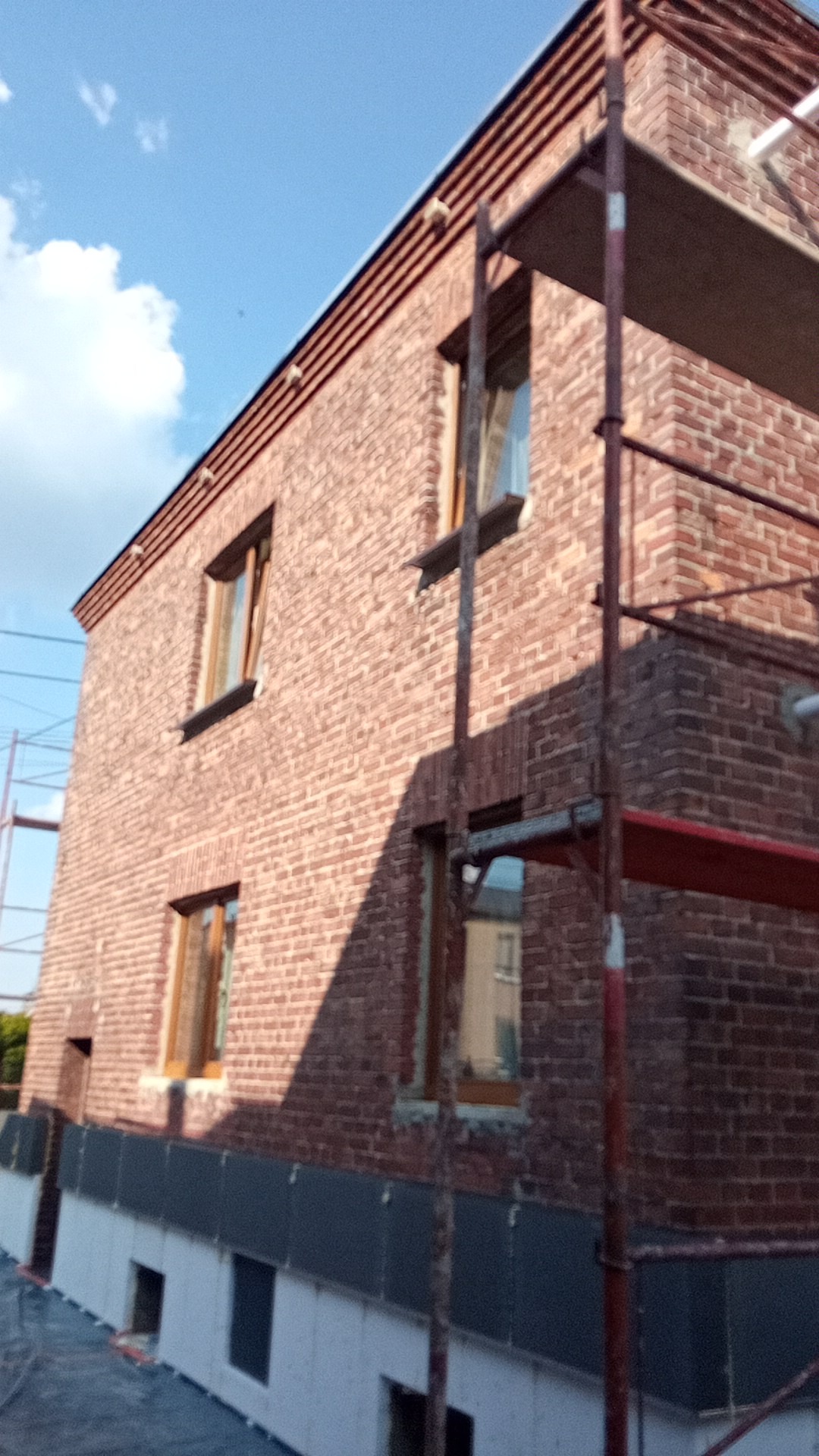 Ceglana elewacja domu w trakcie prac, widoczne rusztowania i fragment ocieplenia fundamentów, błękitne niebo w tle.