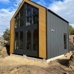 Nowoczesny dom modułowy z dużymi przeszkleniami, okładziną z drewna i szarą elewacją, posadowiony na betonowej płycie, widoczne prace ziemne wokół budynku.