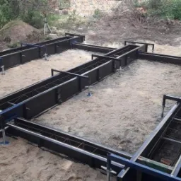 Budowa fundamentów: pracownik w czerwonej koszulce montuje szalunek z metalowymi wzmocnieniami na piaszczystym gruncie.