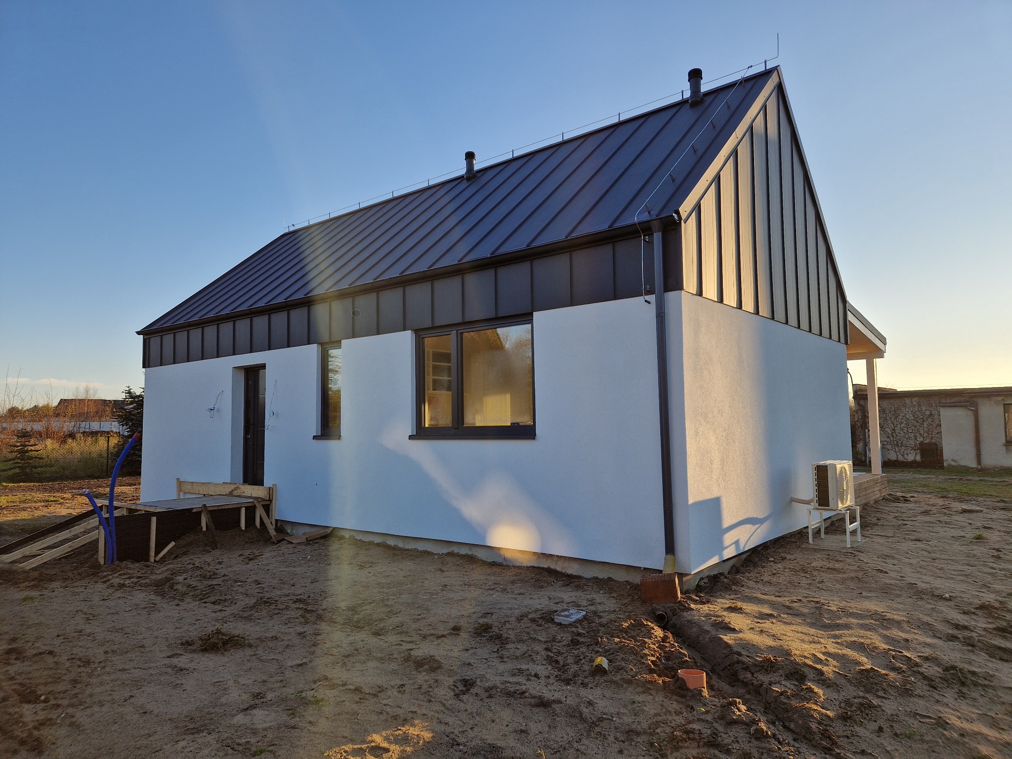 Niedokończony biały dom z czarnym dachem, widoczne elementy instalacji na zewnątrz i przygotowane wejście, otoczenie w stanie surowym.