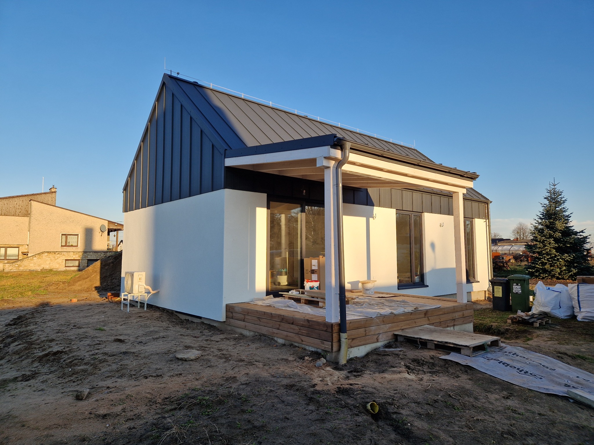 Nowoczesny dom z ciemnym dachem i jasną elewacją, widoczny taras w trakcie wykańczania i otoczenie placu budowy, w tle inne budynki i świerk.