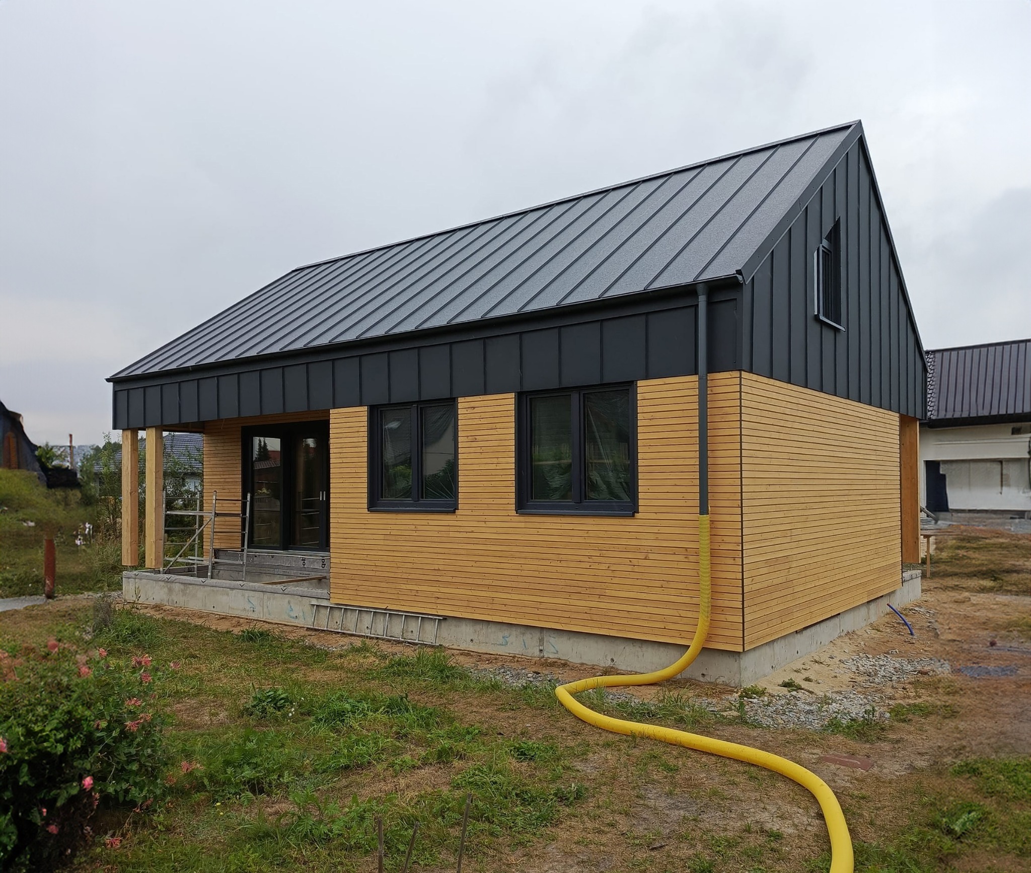 Niedokończony dom z drewnianą elewacją i ciemnoszarą blachą na dachu, widoczne rusztowania i żółty wąż na trawniku.