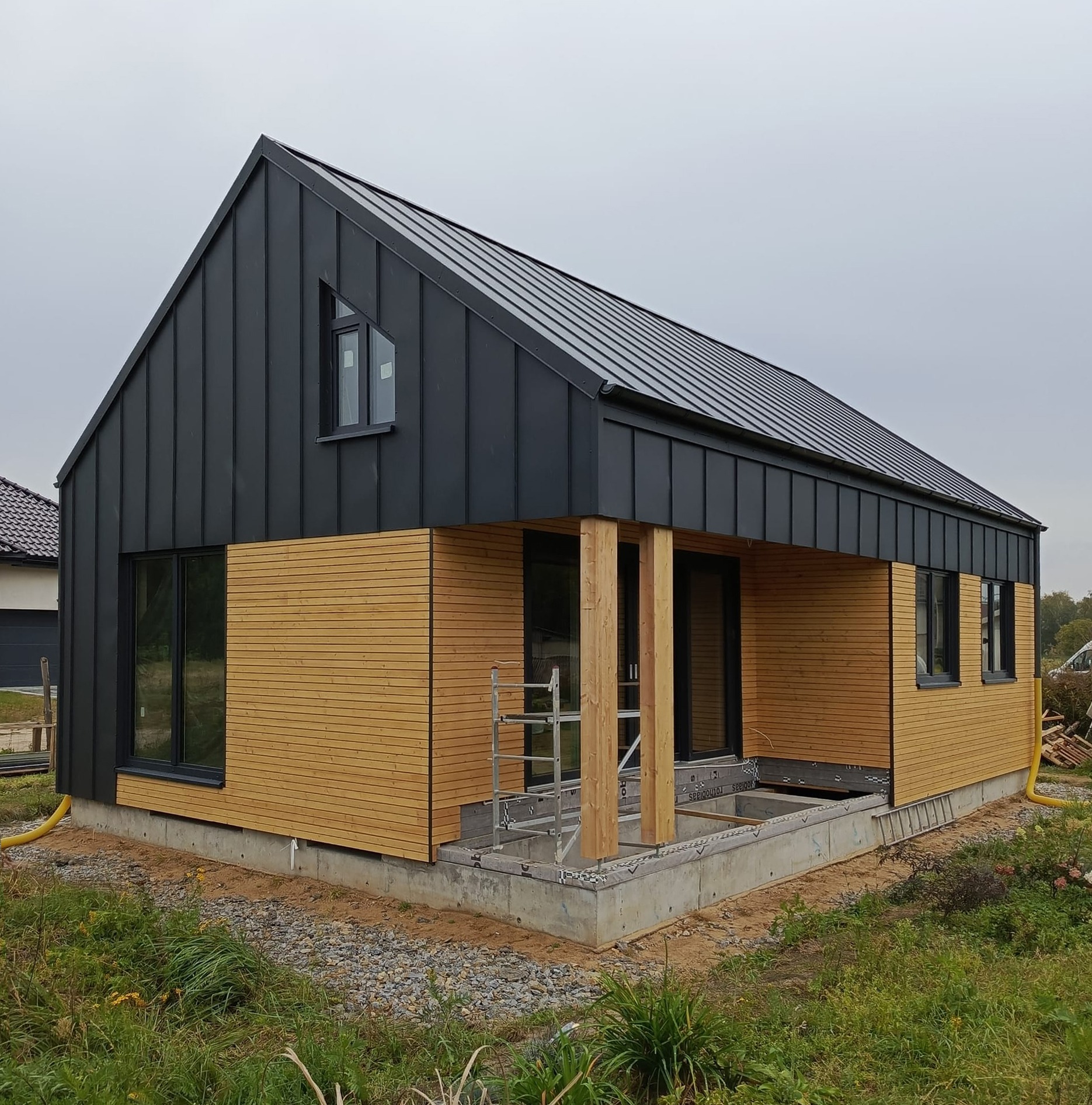 Niedokończony dom z czarną, metalową elewacją i drewnianą boazerią, widoczne rusztowanie i fundament, w tle trawa i inny dom.