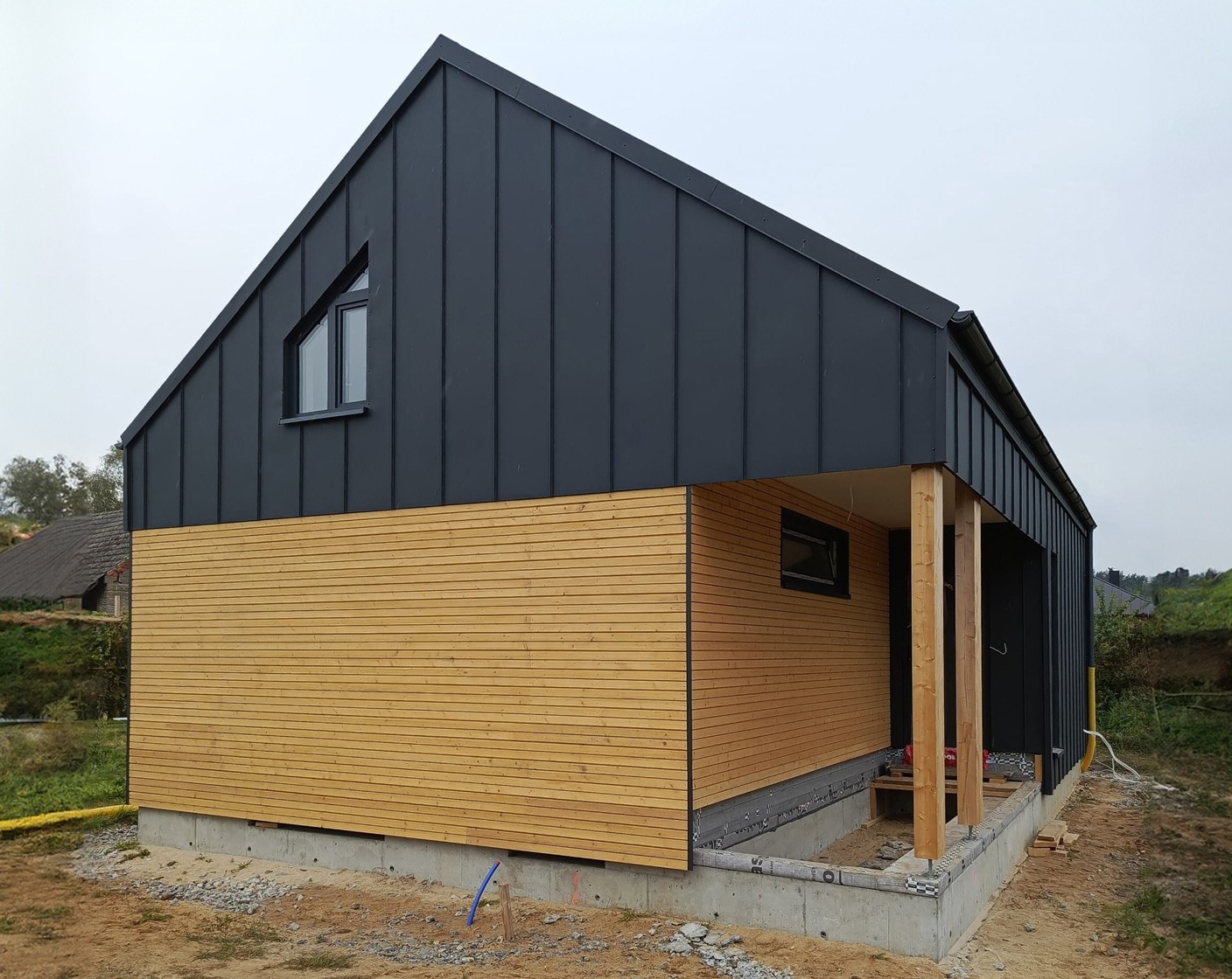Nowoczesny dom z ciemną blachą na górnej części i jasnym drewnem na dolnej, widoczne drewniane słupy werandy i małe okno trójkątne na poddaszu.