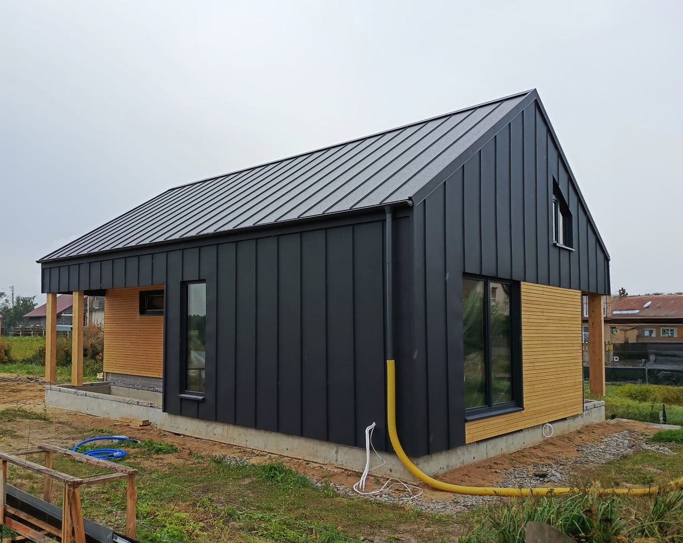 Nowoczesny dom szkieletowy w trakcie budowy, z czarną blachą na elewacji i jasnym drewnem, widoczne niebieskie węże na trawie i żółty wąż wentylacyjny.
