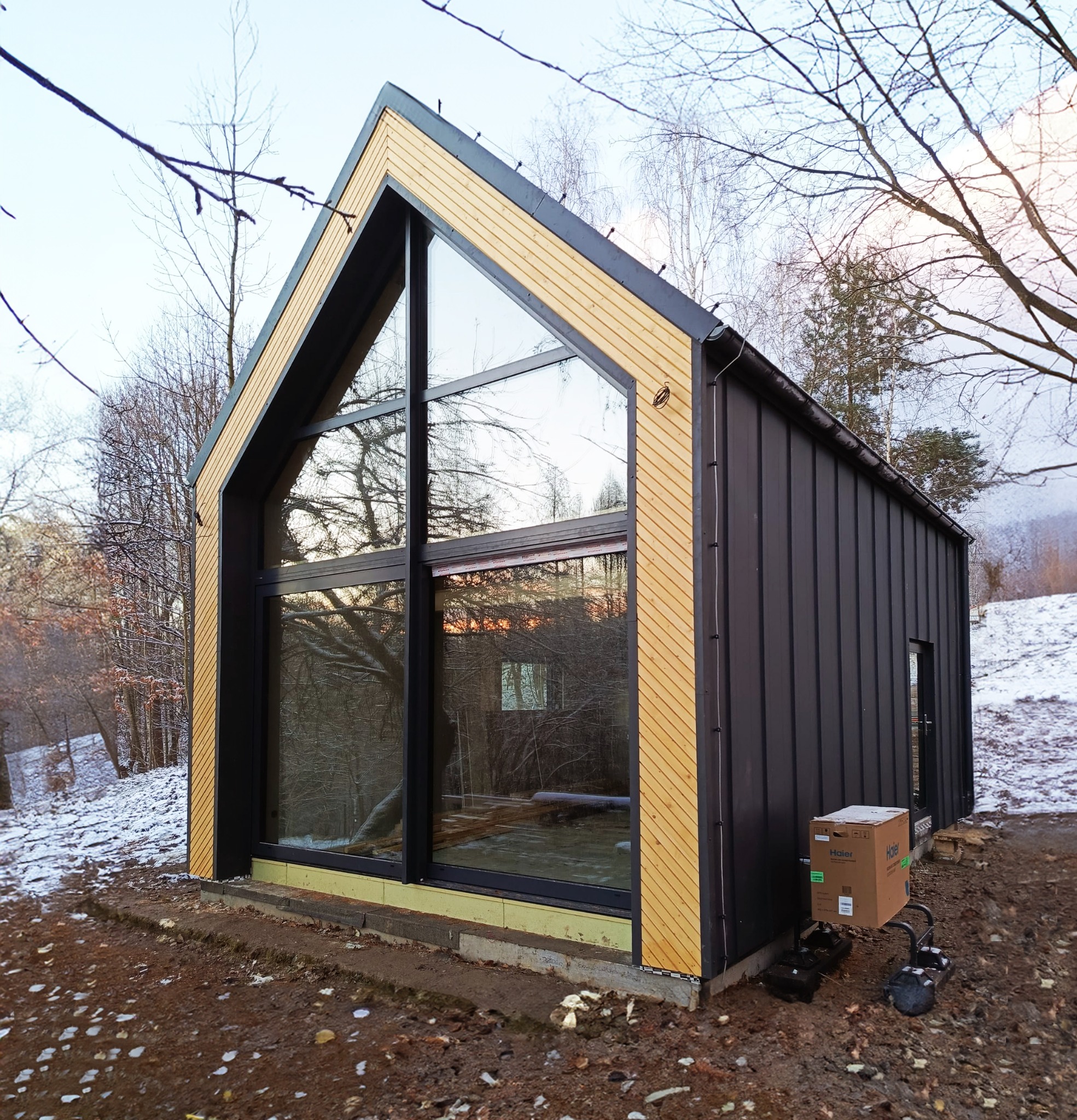 Nowoczesny, czarno-drewniany dom z dużymi oknami, częściowo wykończony, z kartonem i elementami montażowymi na zewnątrz w zimowym krajobrazie.