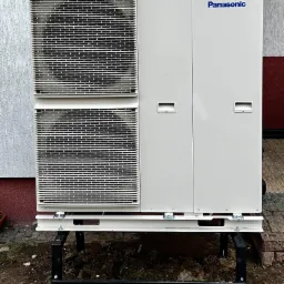 Zewnętrzna jednostka dwuwentylatorowa, marki Panasonic, zamontowana na metalowej konstrukcji przy ścianie budynku z widocznymi elementami instalacji.