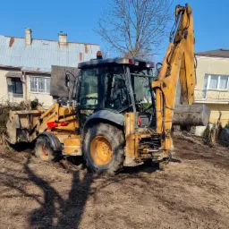 Żółta koparko-ładowarka na nierównym, brudnym terenie, z fragmentami korzeni i gałęzi, w tle domy jednorodzinne pod błękitnym niebem.