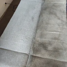 Widok z góry na jasnoszarą kanapę, po lewej stronie widoczna wyraźna różnica w czystości tapicerki po praniu, prawa strona nadal zabrudzona.