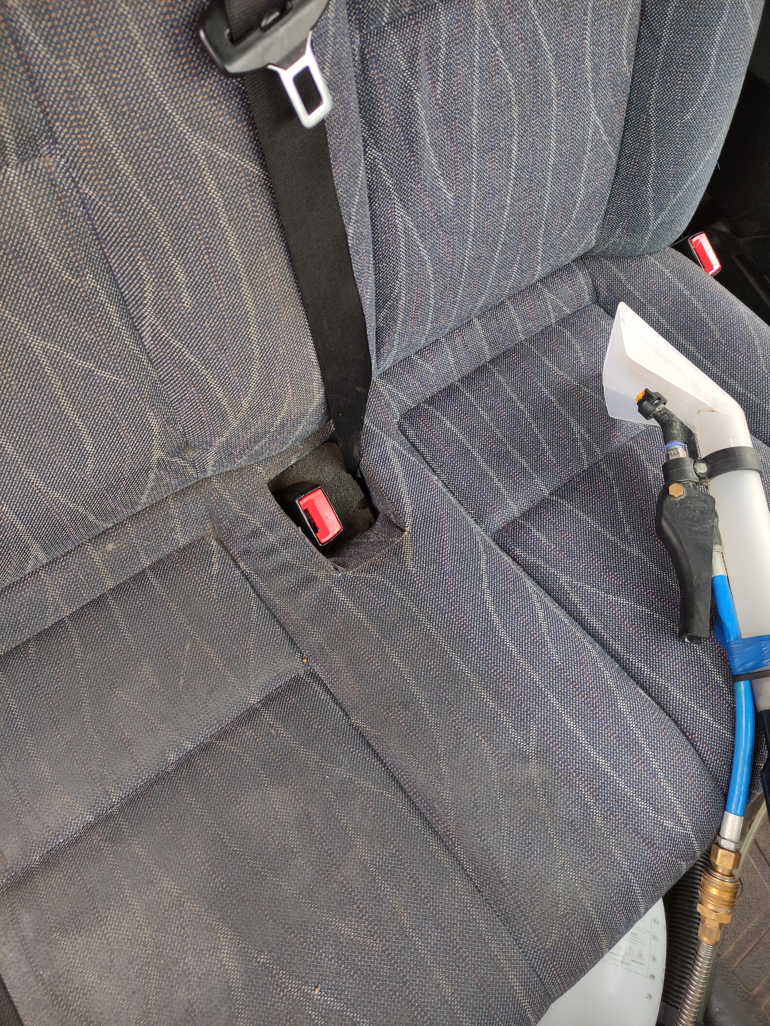 Czyszczenie tapicerki samochodowej na tylnym siedzeniu, widoczny pas bezpieczeństwa i dysza odkurzacza piorącego z niebieskim wężem.