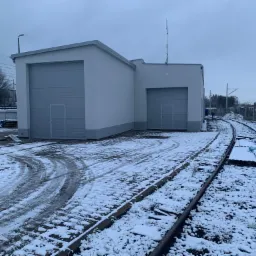 Biały budynek z dwoma bramami garażowymi, zlokalizowany przy torach kolejowych pokrytych śniegiem w pochmurny dzień.