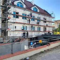 Ekipa budowlana na rusztowaniu podczas prac wykończeniowych elewacji trzypiętrowego budynku mieszkalnego z poddaszem, okna zabezpieczone folią, widoczne materiały budowlane i tablica ostrzegawcza.