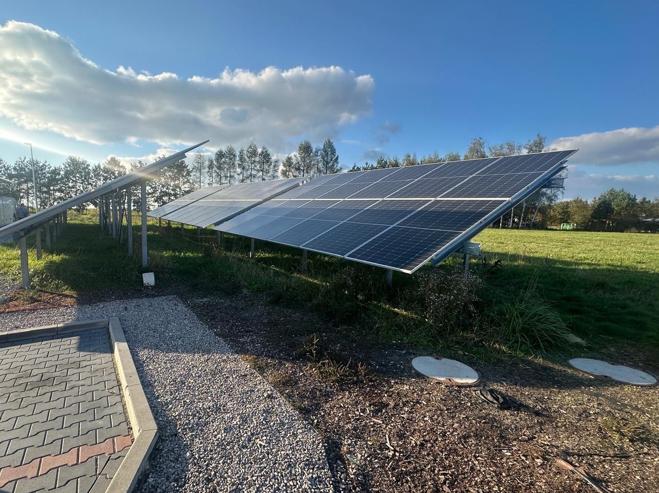 Rząd paneli słonecznych na łące, z boku widoczny chodnik z kostki. Błękitne niebo z białymi chmurami w tle. Widoczne trawy i drzewa w oddali.