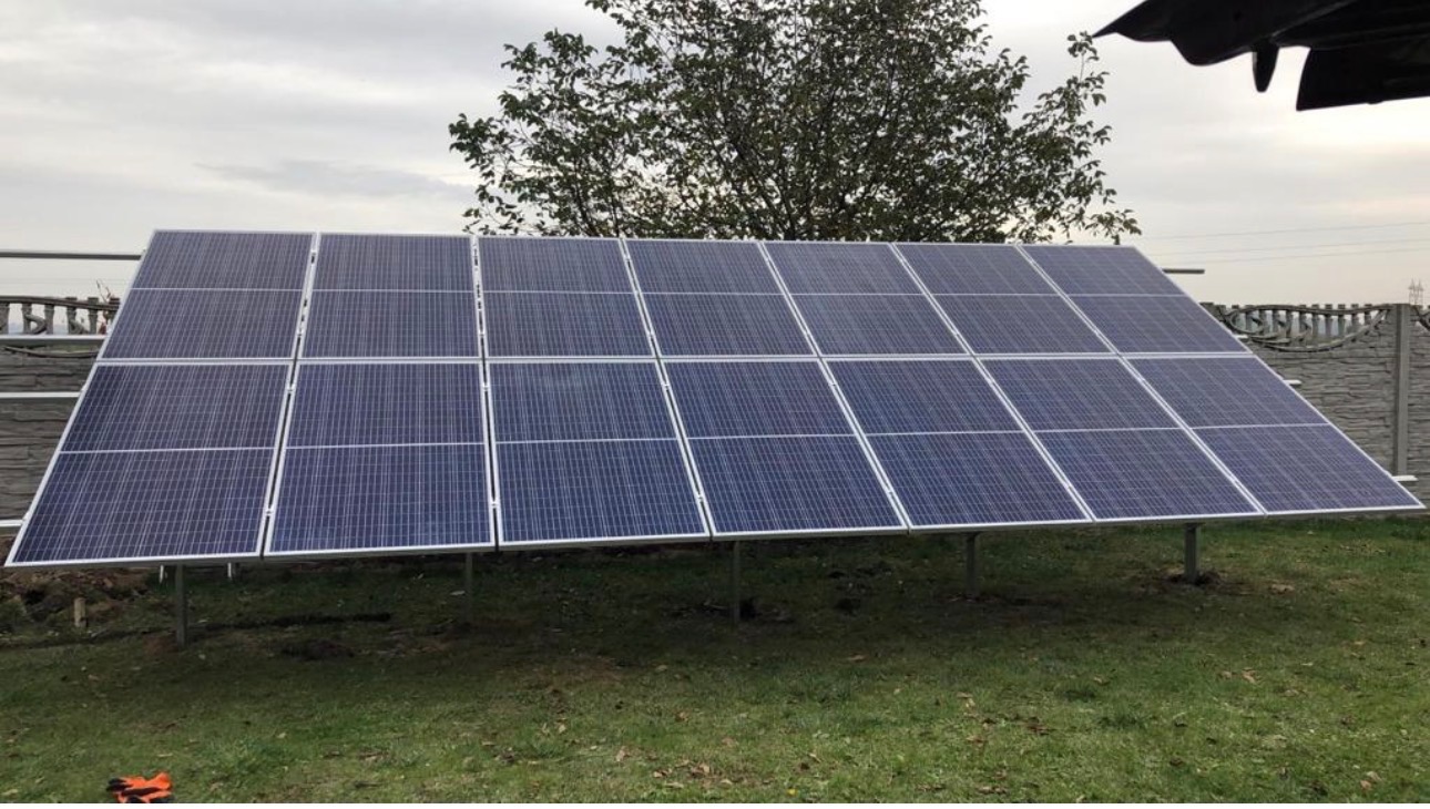 Instalacja fotowoltaiczna na trawniku, widok z boku. Panele ustawione pod kątem, szara pogoda, w tle drzewo i ogrodzenie z betonowych elementów.