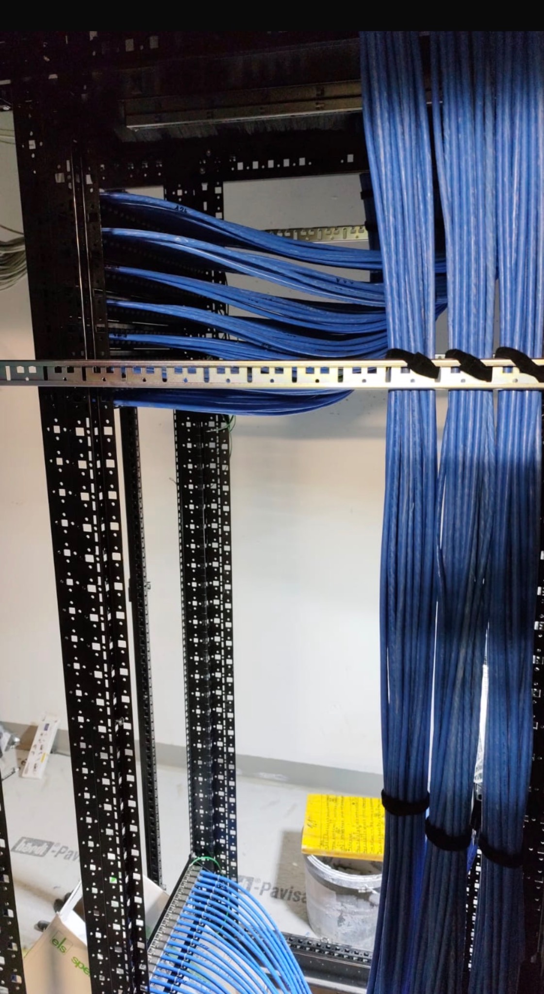Uporządkowane niebieskie kable w szafie rack, zabezpieczone czarnymi opaskami. Widoczne metalowe elementy konstrukcyjne i fragment podłogi z wiadrem.