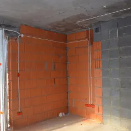 Surowy stan wnętrza z widoczną instalacją elektryczną: przewody poprowadzone po ścianach z cegły ceramicznej i bloczków betonowych, puszki instalacyjne zamontowane w ścianie, widoczne okno...