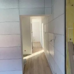 Wnętrze w trakcie remontu: ściany wyłożone jasnymi panelami z delikatną fakturą, widoczne przewody elektryczne wystające ze ściany, podłoga wyłożona panelami drewnopodobnymi, w tle widoczne kolejne...