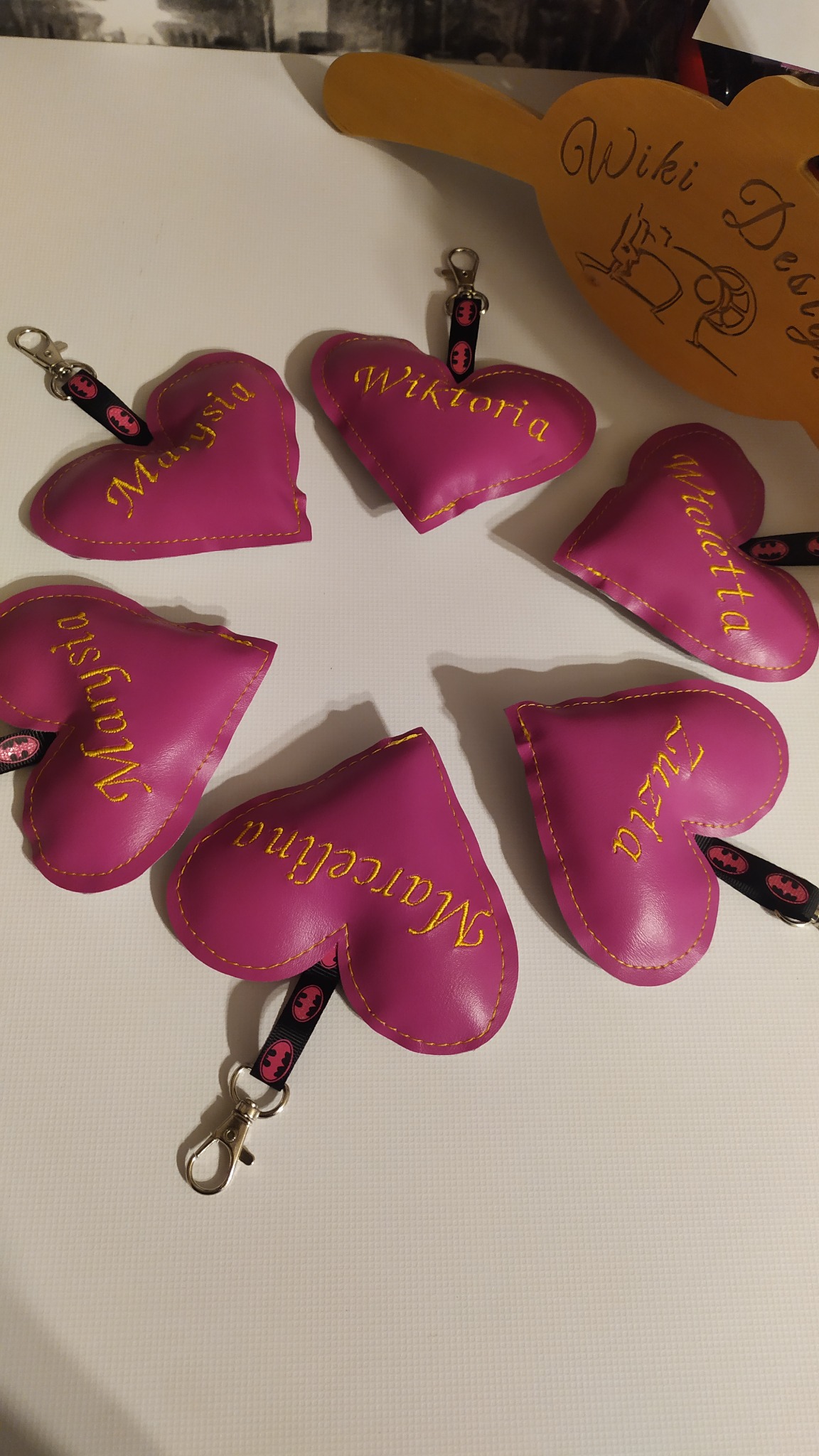 Różowe serca z imionami wyszytymi żółtą nicią, przymocowane do breloczków z motywem Batmana, ułożone na białym tle, obok drewnianego przedmiotu z napisem 'Wiki Design'.