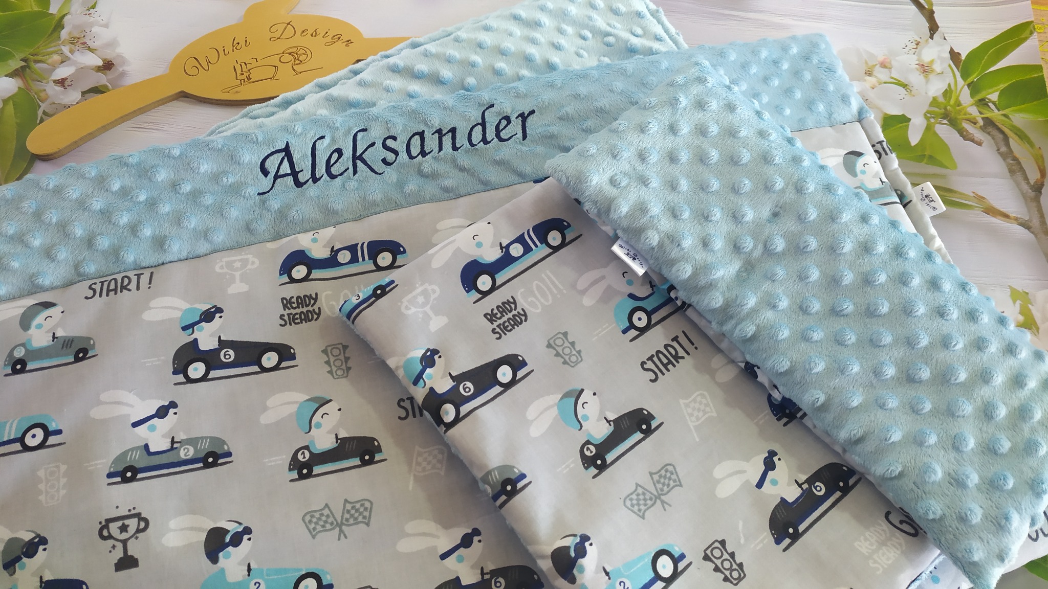 Komplet dziecięcej pościeli z motywem wyścigów samochodowych i królików, błękitna kołderka z imieniem 'Aleksander', drewniany wieszak 'Wiki Design', całość na jasnym drewnianym tle z gałązką...